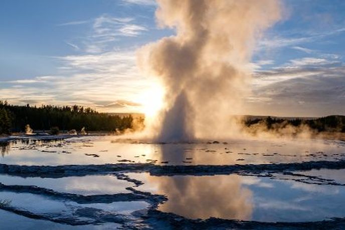 Steam erupts from a geothermal geyser on a snowy landscape at sunrise, with a calm river and forest in the background