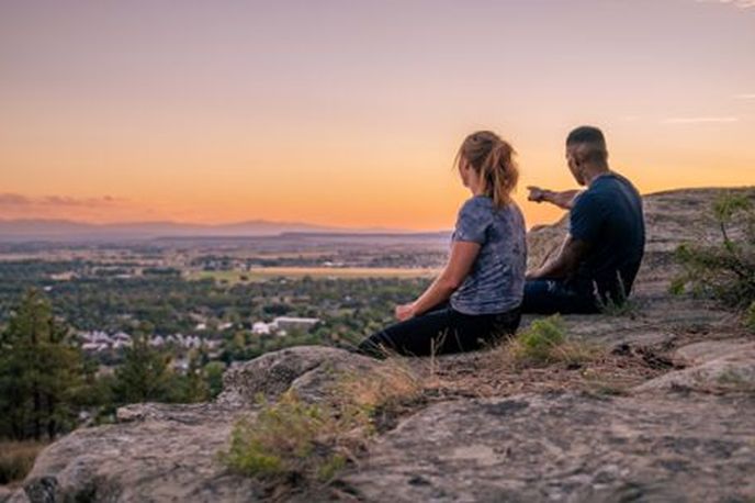 Two people sitting on a rocky overlook at sunset, looking over a Montana town and valley