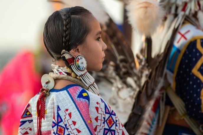 Native American woman in traditional regalia wearing beadwork and earrings, surrounded by other regalia at a cultural gathering in Montana