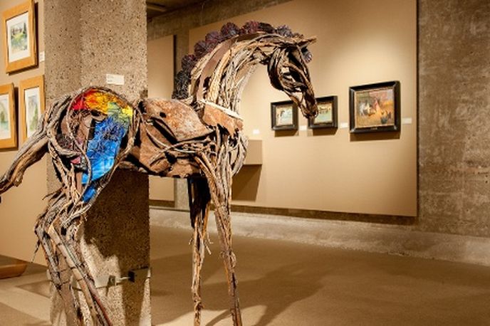 Wooden horse sculpture with mounted rider in an indoor gallery space with framed artwork on the walls