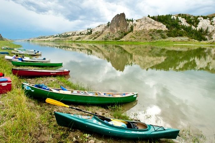 Green and red kayaks on the shore of a river with boats moored nearby, looking toward whitewater rapids in Montana