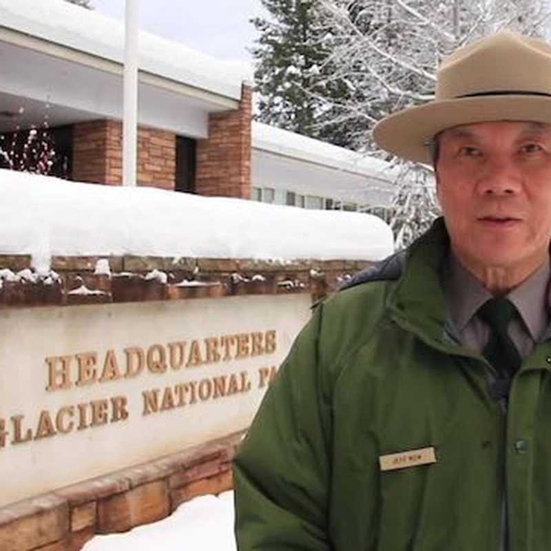 Park visitor wearing a hat and green jacket in front of the “Headquarters, Glacier National Park” sign during snowy weather in Montana