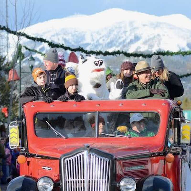 Families ride in an open red vintage truck through a snowy mountain town lined with winter decorations and crowds
