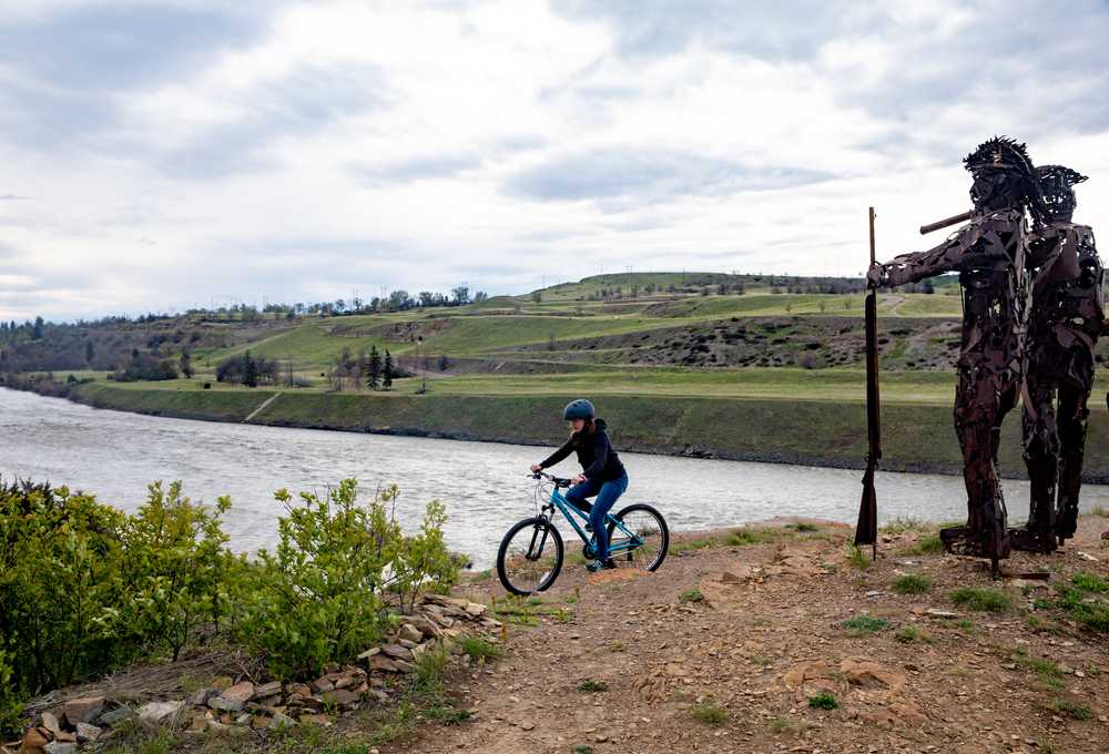 Person riding a blue bicycle along a riverbank in rural Montana with rolling green hills in the background