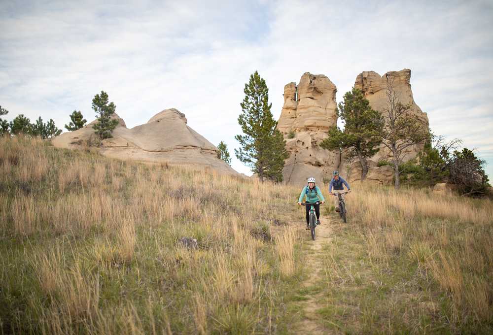 Two hikers walk a trail through tall grass toward red rock formations in Montana