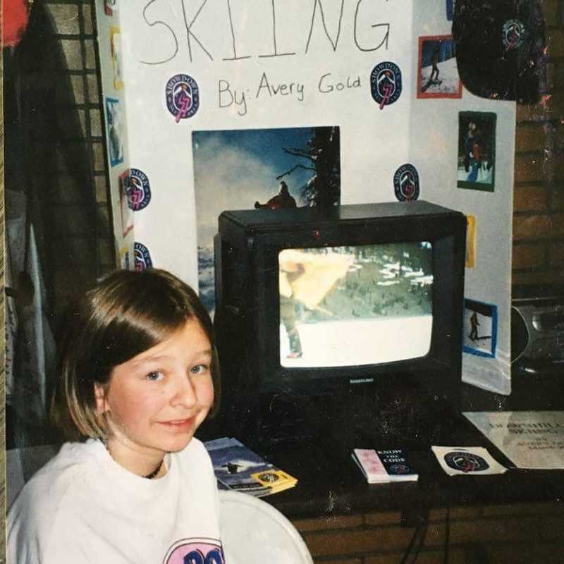 Person seated at a display booth with a “SKIING” banner and “By Avery Gould” sign, promoting skiing memorabilia and information in an indoor venue