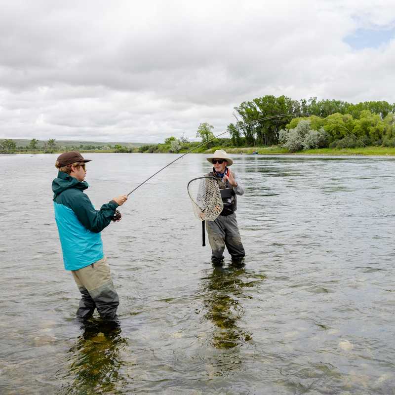 Two anglers wade in the Missouri River while fishing near a grassy shoreline and trees in Montana