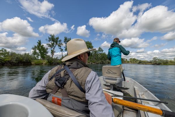 Two anglers in a small fishing boat on a calm Montana river, with one holding a fishing rod and the other seated at the bow under a partly cloudy sky