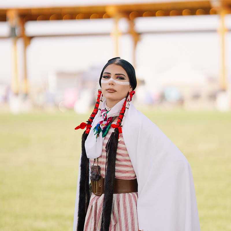 Woman wearing traditional Native American regalia with braided hair and ribboned scarf at an outdoor event in Montana