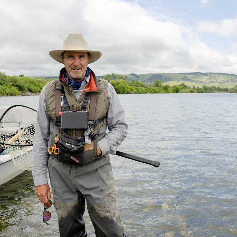 Angler standing on a boat holding a fishing rod on a wide river with Montana hills and mountains in the background