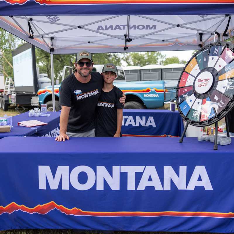 People standing at a Montana tourism booth with a large “MONTANA” banner and a wheel display under a canopy tent