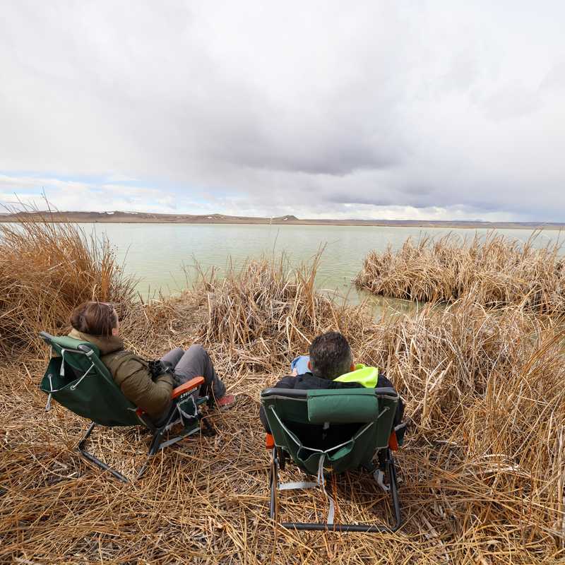 Two people relaxing in folding lawn chairs in a field of tall grass near a lake under cloudy skies in Montana