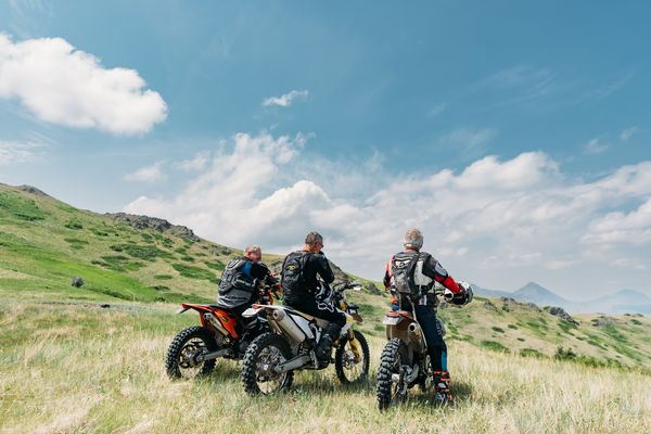 Three riders with adventure motorcycles travel along a grassy mountain trail under a blue sky in Montana