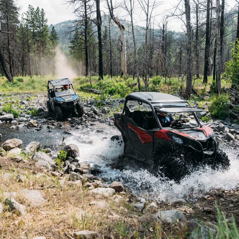 Two off-road vehicles splash through a shallow creek in a forest clearing near mountains in Montana
