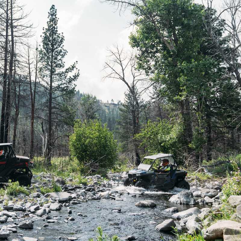 Two off-road vehicles navigate a rocky stream in a forested mountain area, surrounded by sparse trees and lush greenery under a cloudy sky.