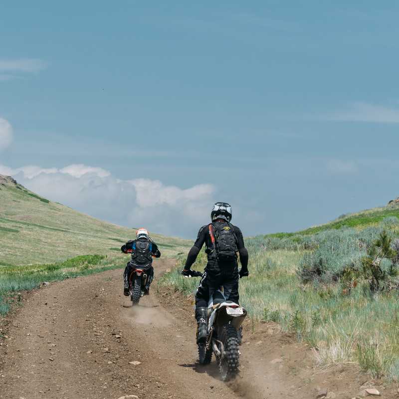 Two cyclists riding on a dirt road through grassy hills with a mountain valley in the distance