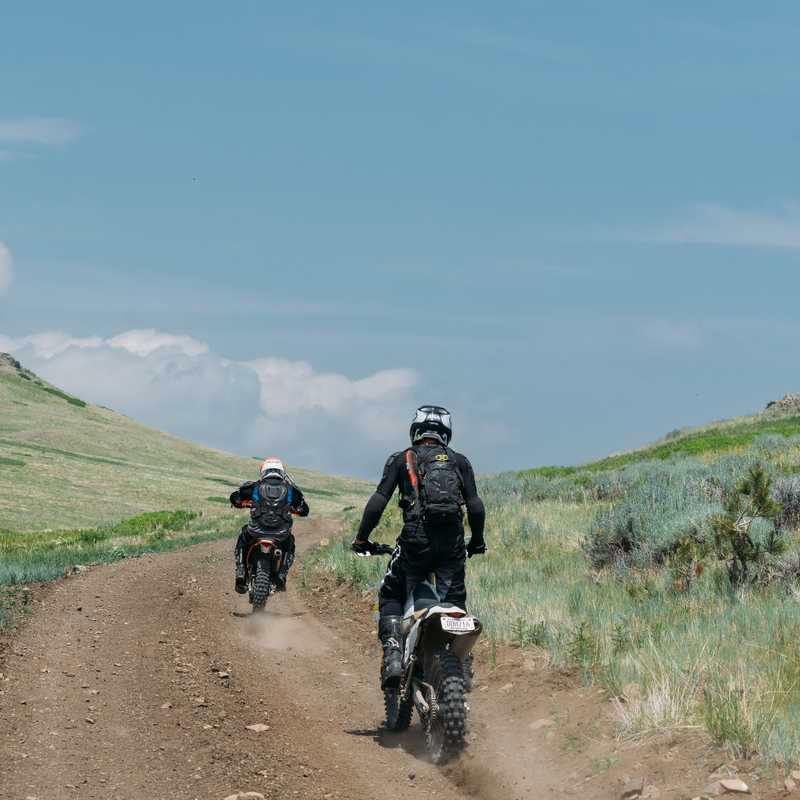 Two cyclists riding on a dirt road through grassy hills with a mountain valley in the distance