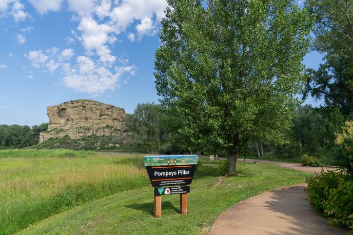 A scenic view of Pompeys Pillar with a grassy field and a path leading by a sign in the foreground.
