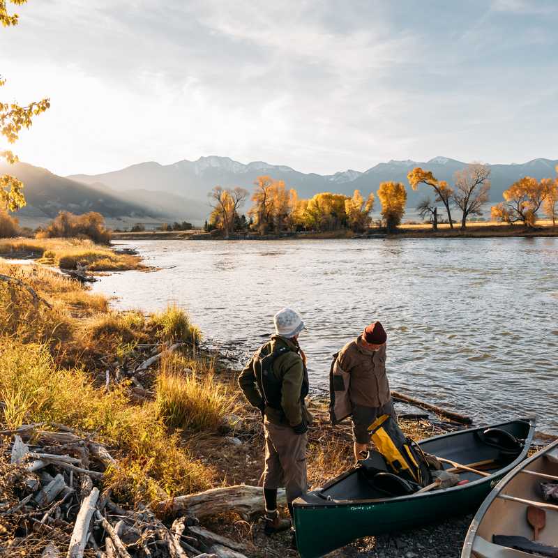 Two people in a small canoe on a Montana river at sunset with autumn trees and distant mountains along the shore