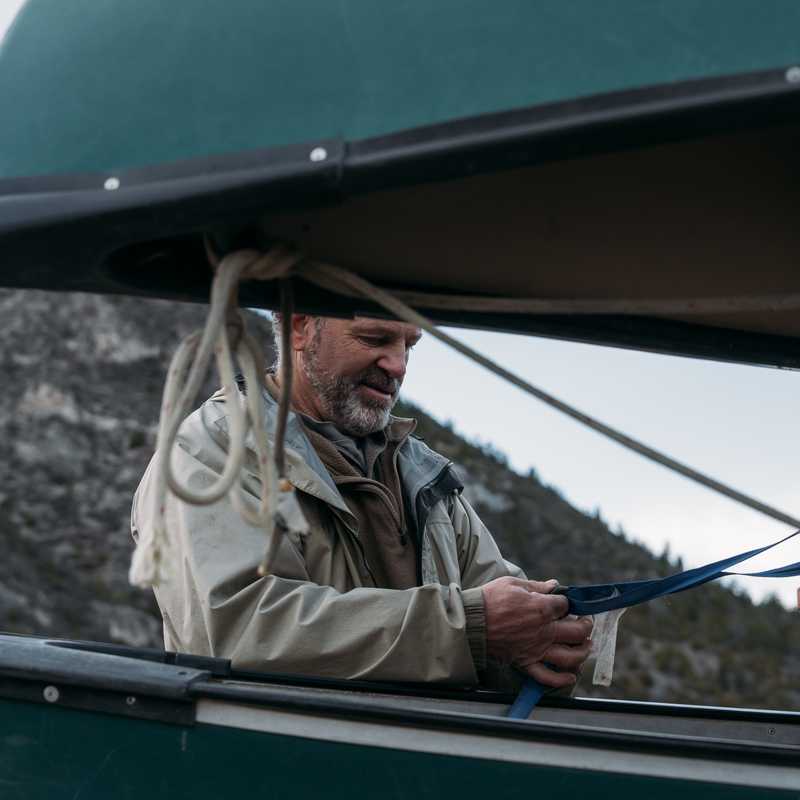 Man adjusting a sailboat rigging on a lake with mountains in the background