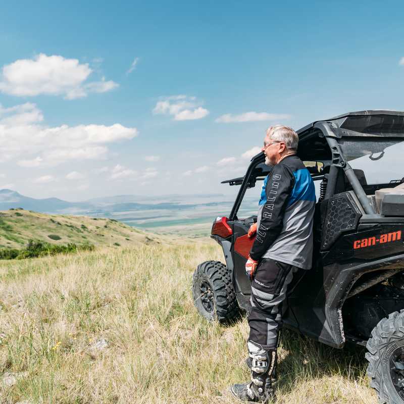 Man standing beside an off-road Can-Am vehicle on a grassy Montana hillside with mountains in the distance