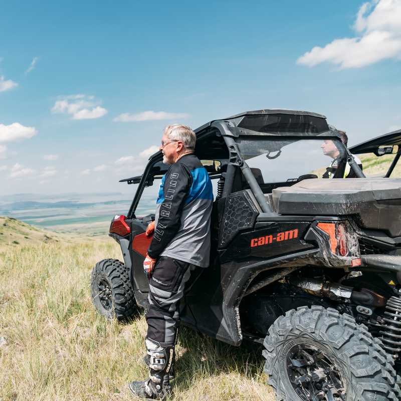 Man standing beside an off-road Can-Am vehicle on a grassy Montana hillside with mountains in the distance