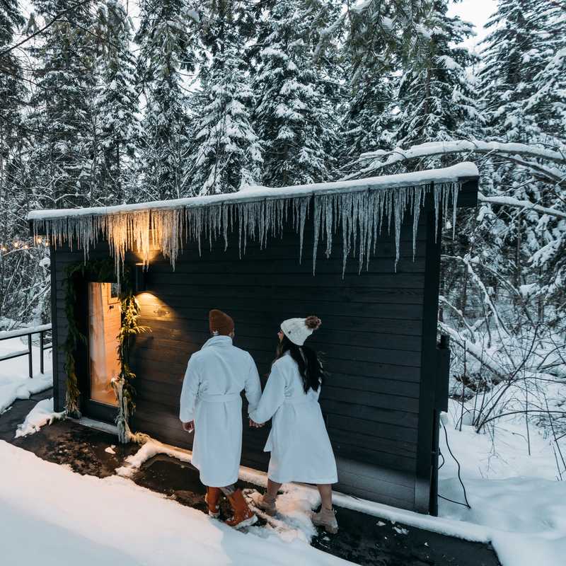People in winter clothing warming at a small wooden sauna with icicles in a snowy forest