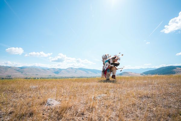Person in colorful regalia dancing on an open grassland with mountain views under a blue sky