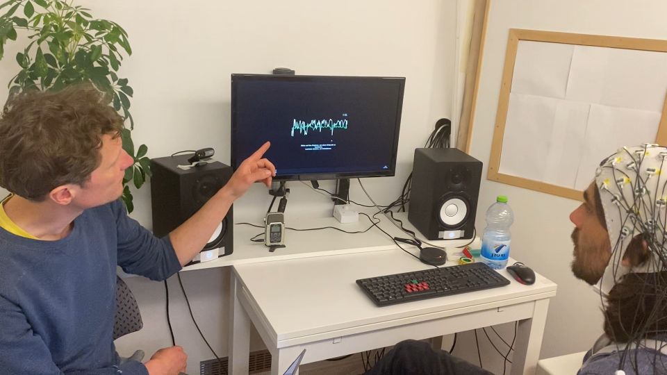 Two people in a room with one pointing at a monitor displaying waveforms. The other wears a cap with sensors. Speakers and a keyboard are on the desk.