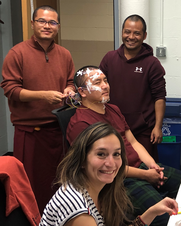 A person with electrodes on their face sits smiling, surrounded by three others, including one holding wires, in a room with a relaxed atmosphere.