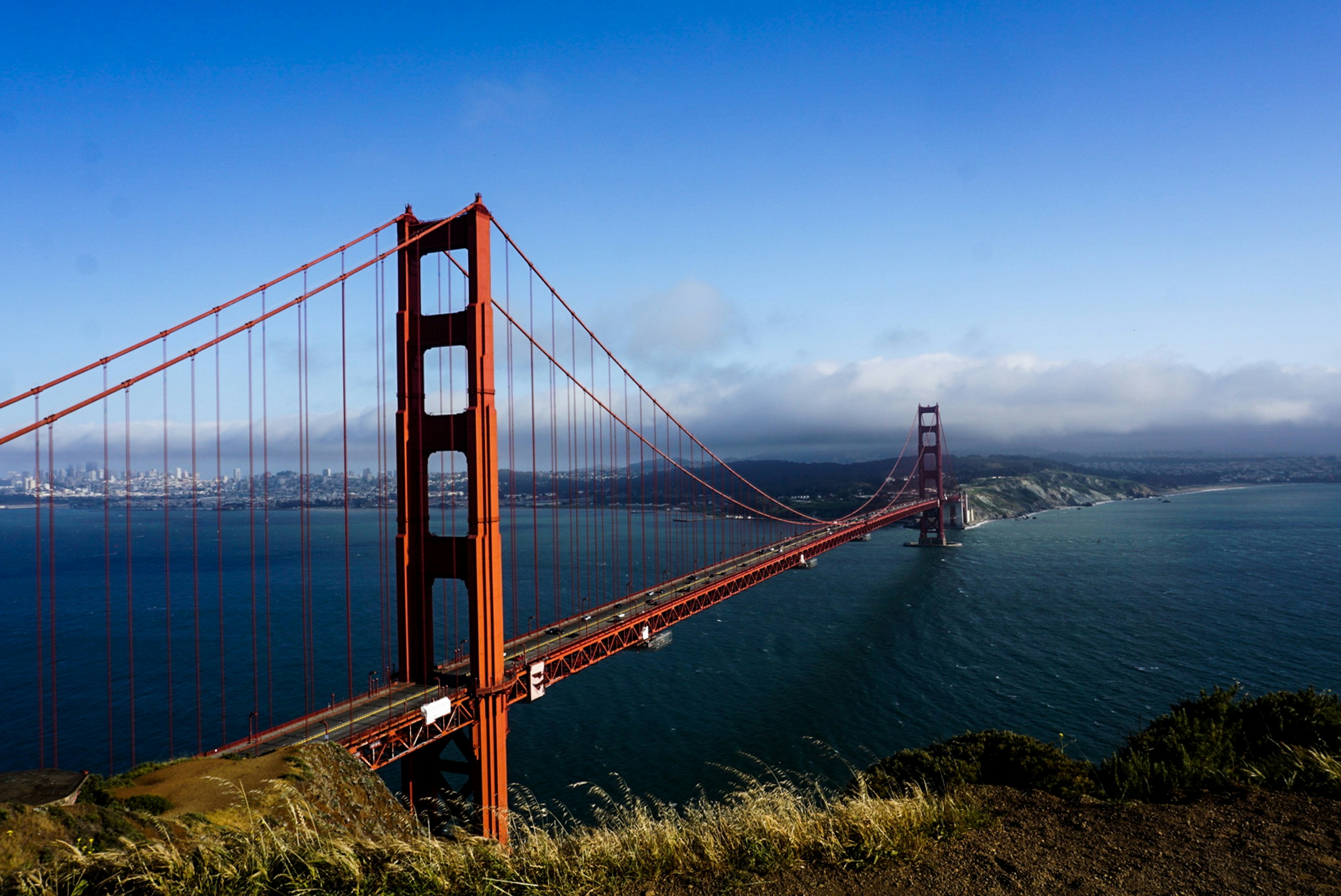 Golden Gate Bridge spanning blue waters, with San Francisco skyline in the background under a clear blue sky.