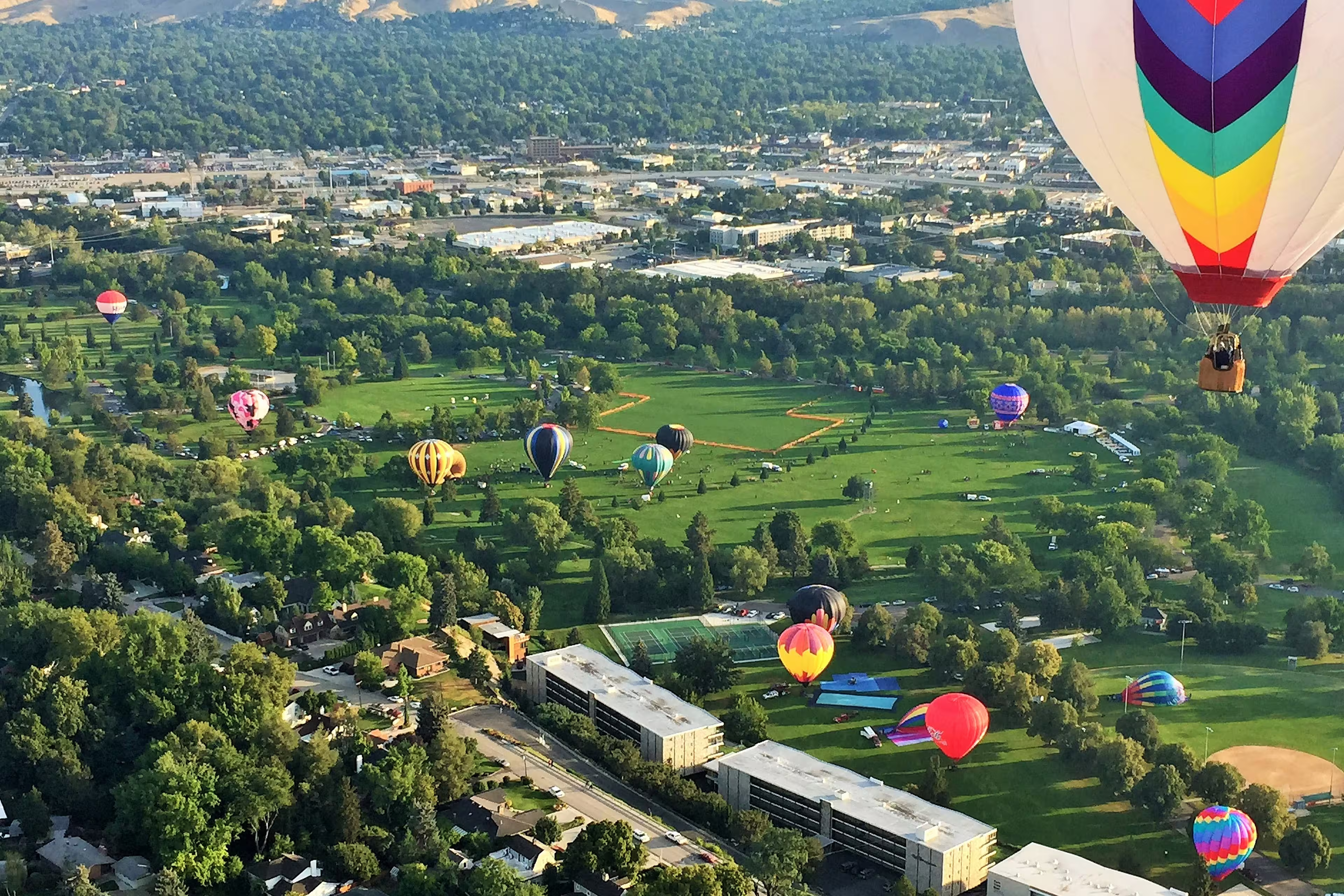 Aerial view of colorful hot air balloons floating over a lush green park in Boise, Idaho, with a cityscape and hills in the background.