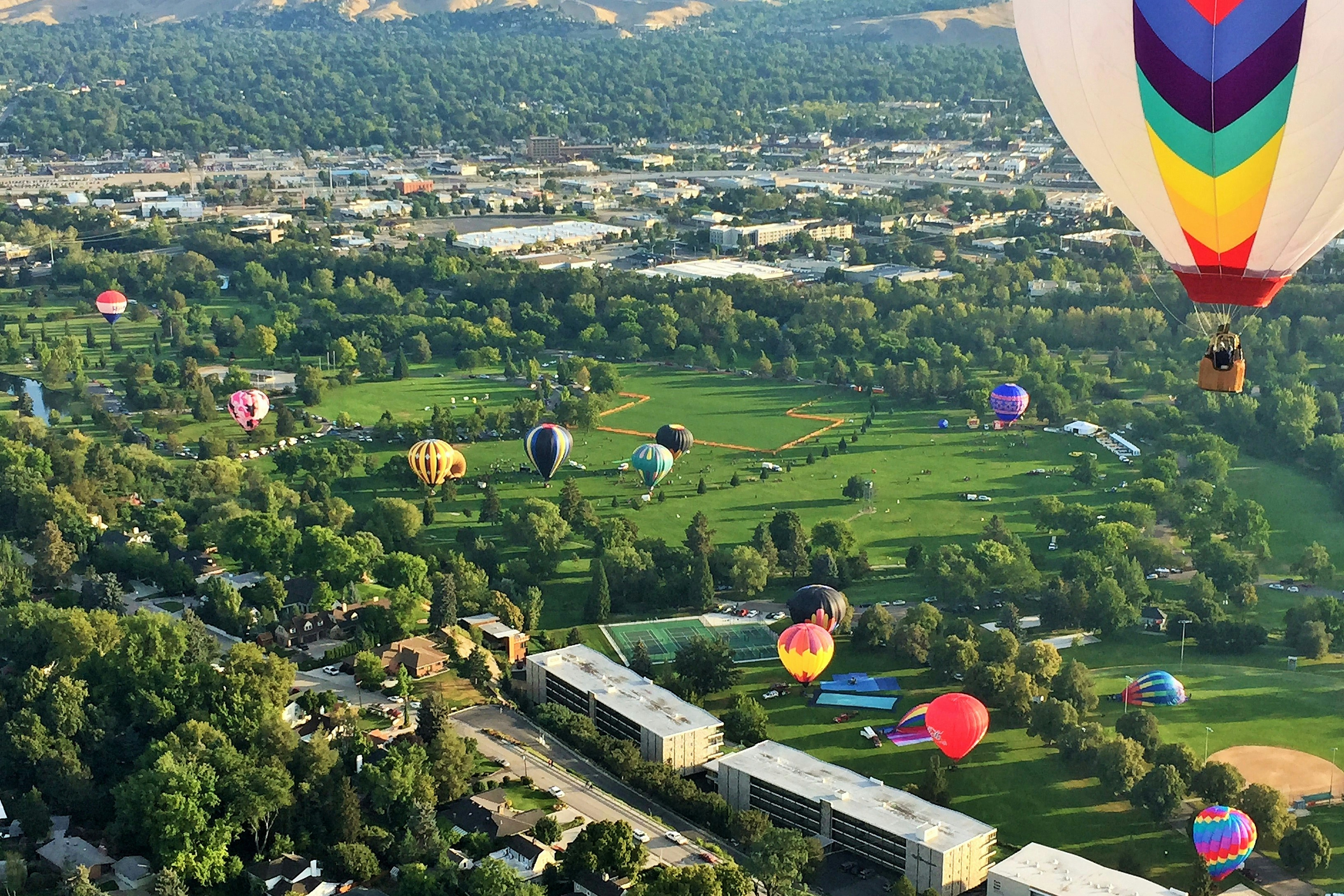 Aerial view of colorful hot air balloons floating over a lush green park in Boise, Idaho, with a cityscape and hills in the background.
