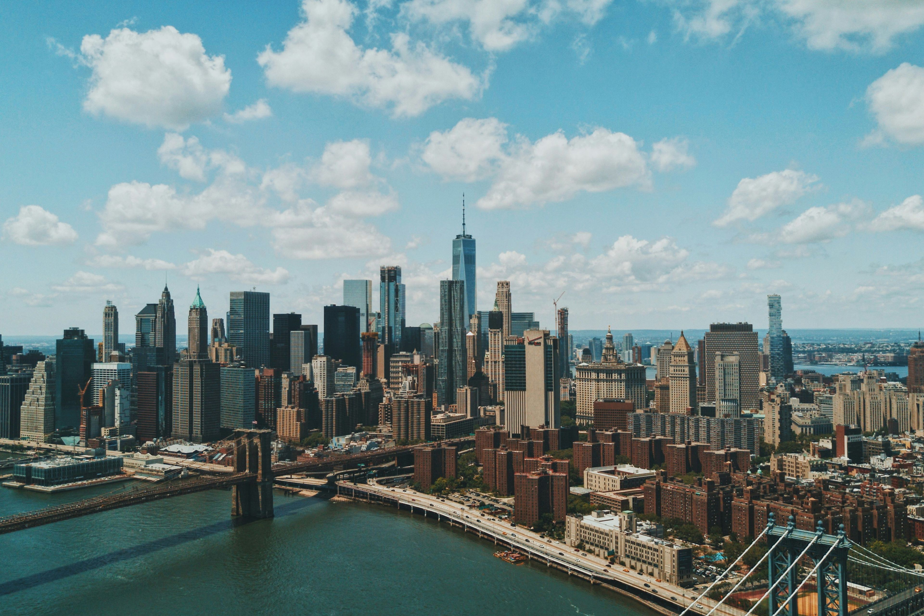 Aerial view of New York's city skyline with skyscrapers, a bridge, and a river under a partly cloudy sky.