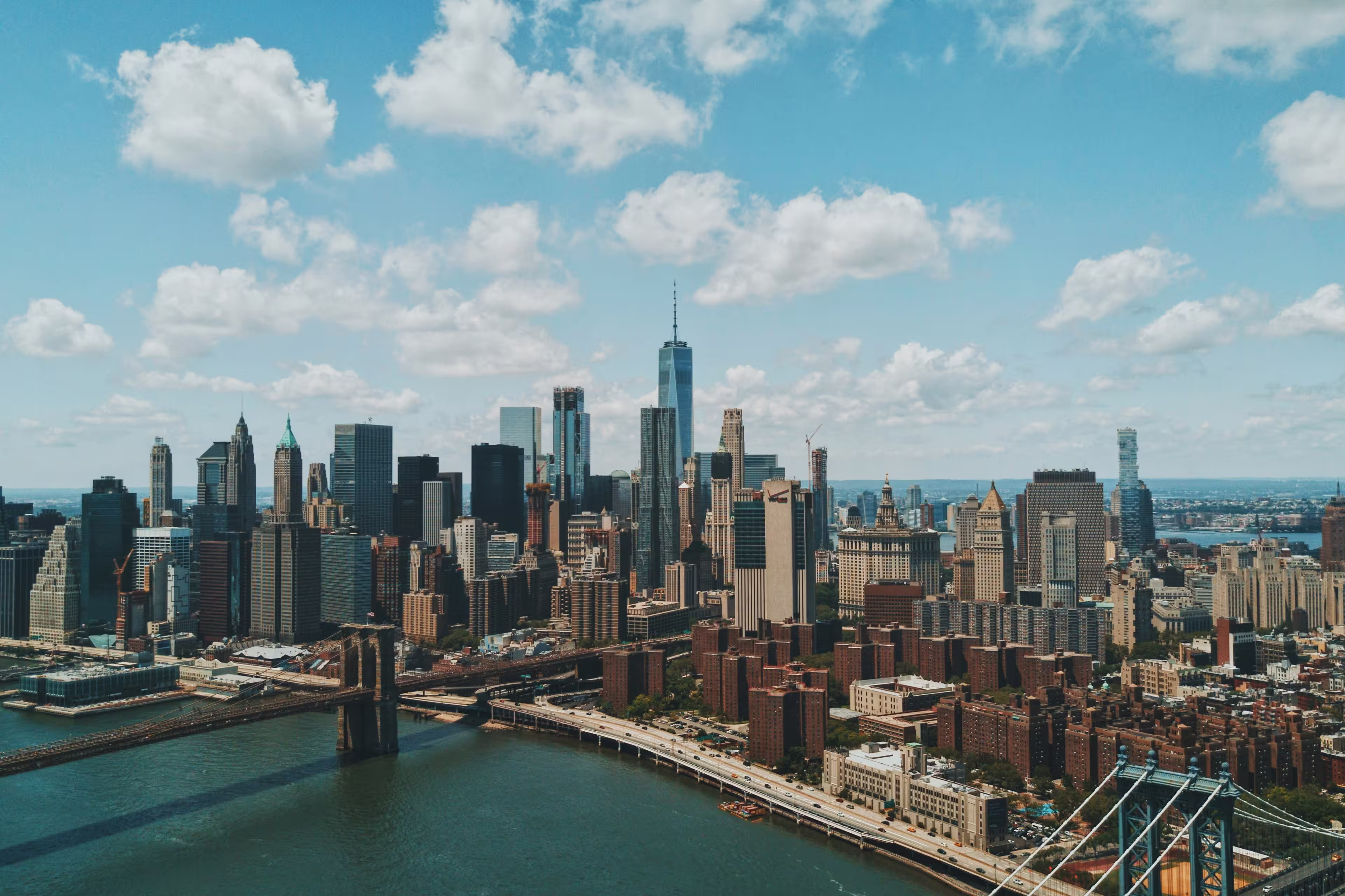 Aerial view of New York's city skyline with skyscrapers, a bridge, and a river under a partly cloudy sky.