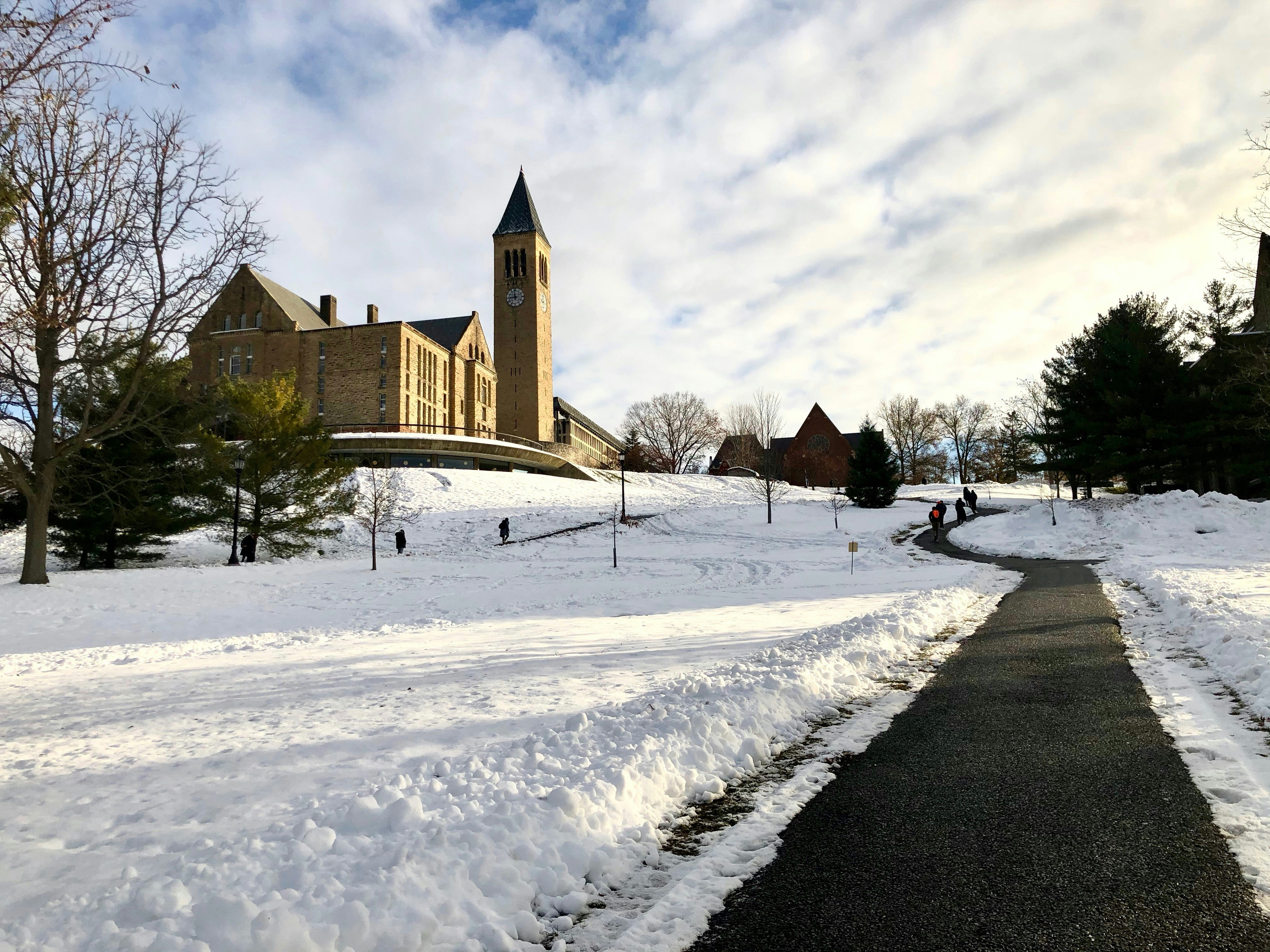 Snow-covered landscape in Ithaca, New York, with a historic building featuring a clock tower, a winding path, and people walking under a partly cloudy sky.