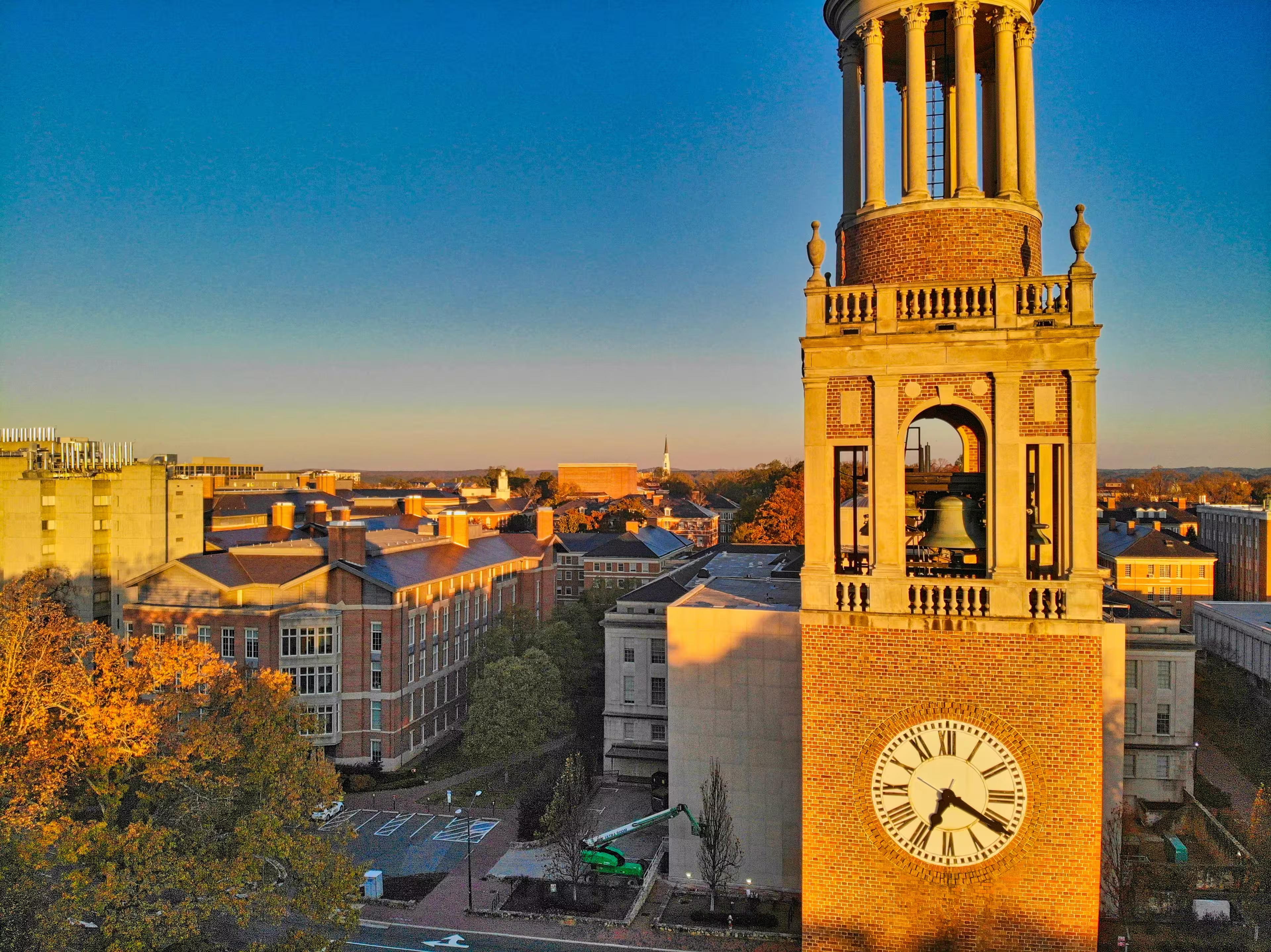 Clock tower overlooking a university campus in Chappel Hill, North Carolina, with red-brick buildings and autumn trees under a clear blue sky at sunset.