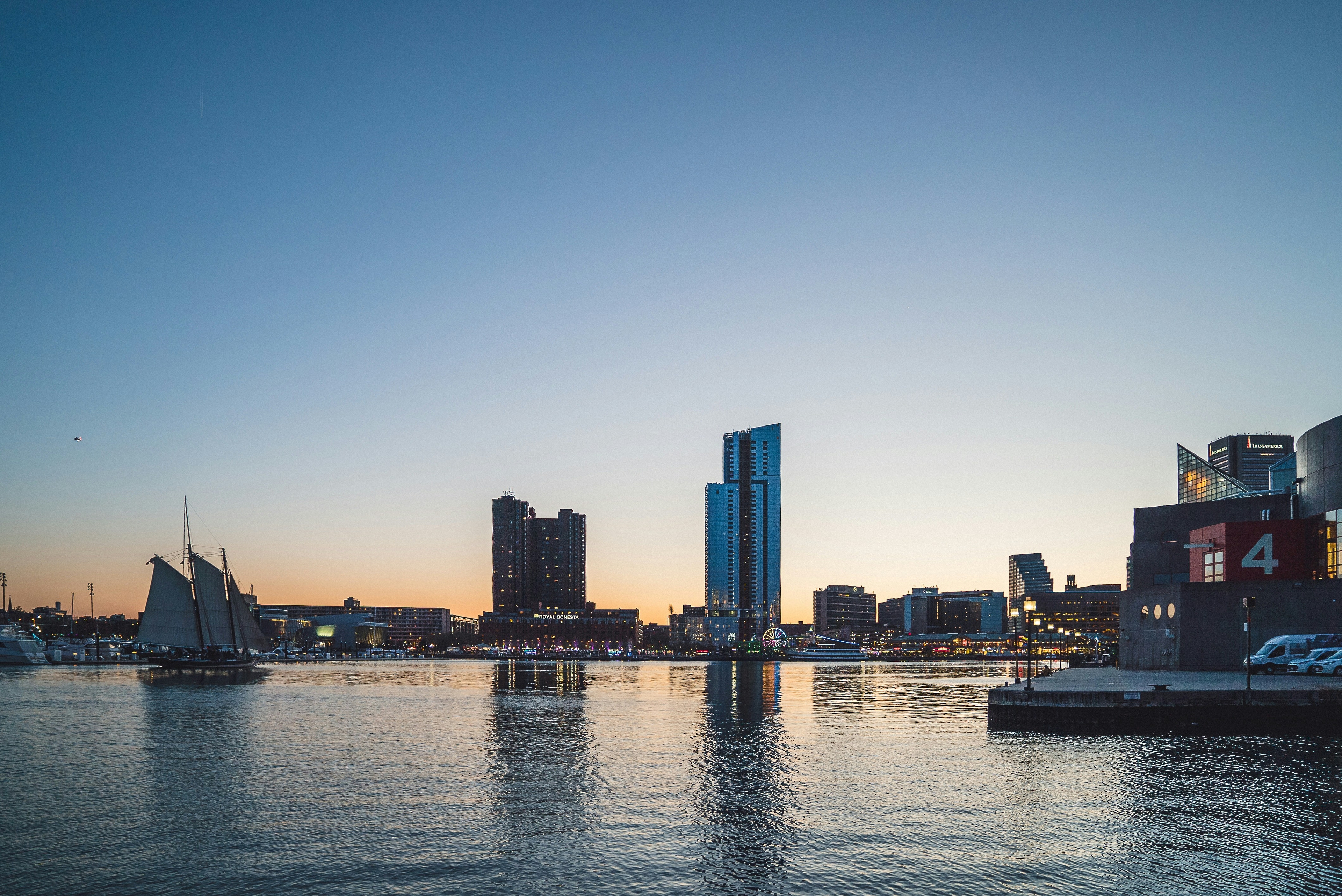 Baltimore's skyline at dusk with tall buildings, a sailing ship on calm water.