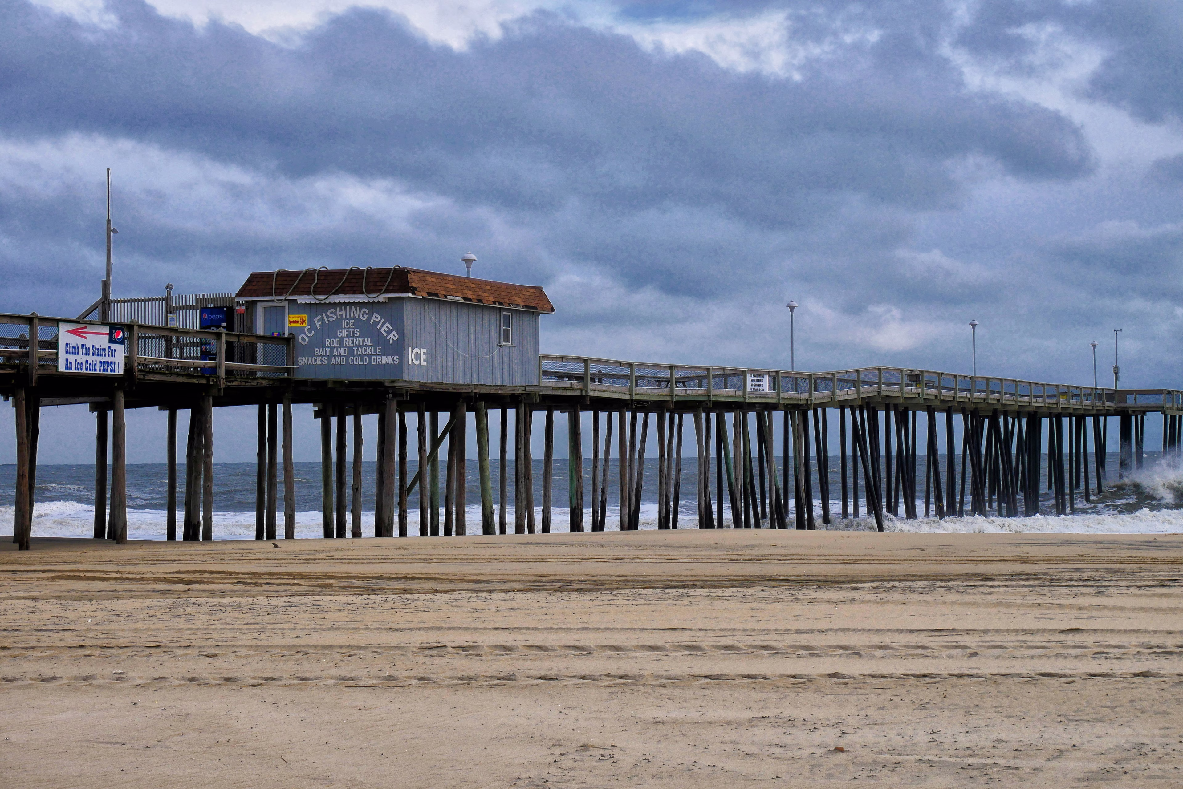 Ocean City fishing pier extends over the ocean under a cloudy sky, with waves crashing and a sandy beach.