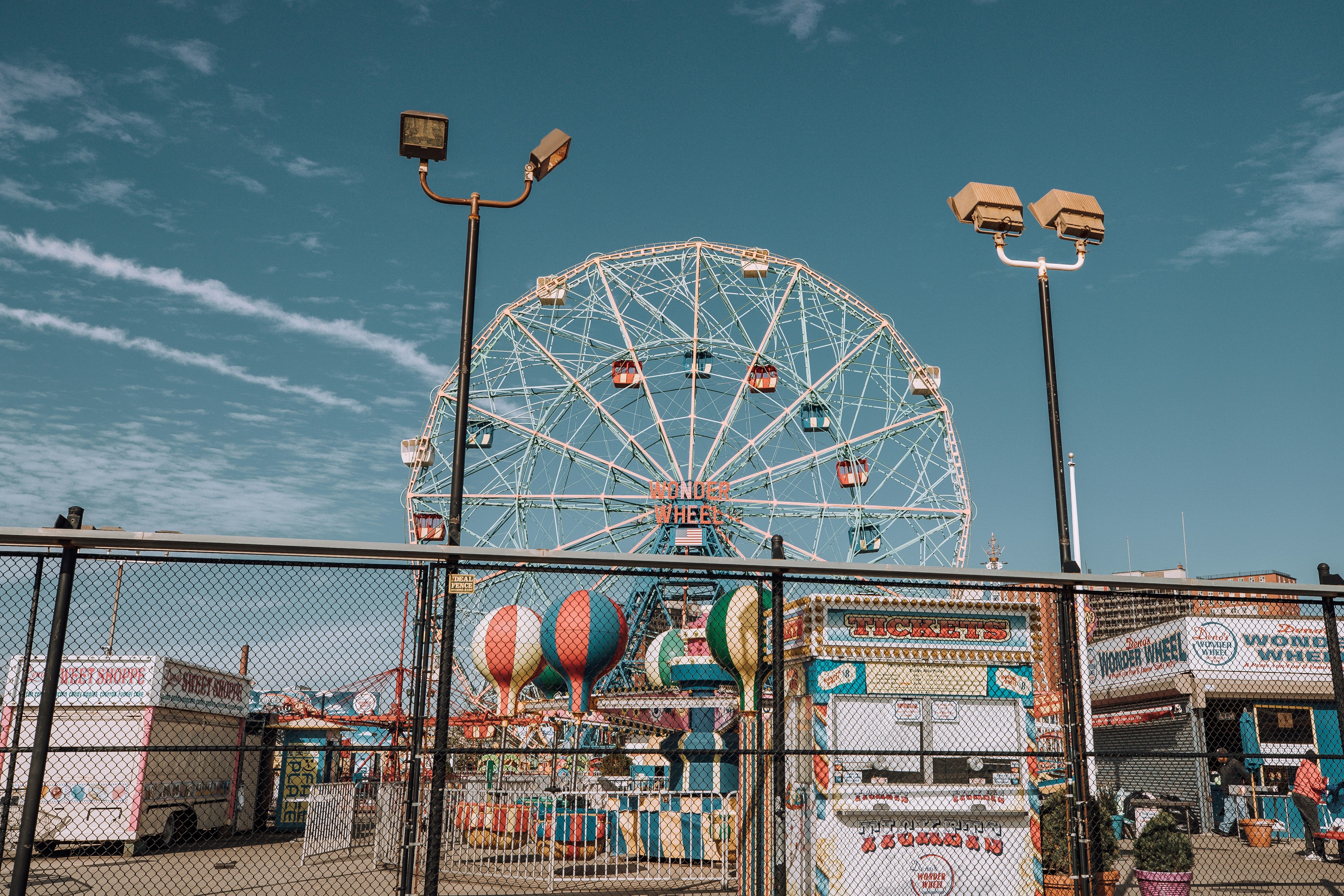 View of the Coney Island Ferris wheel on a clear, bright day with blue skies and a foreground filled with fairground stalls.