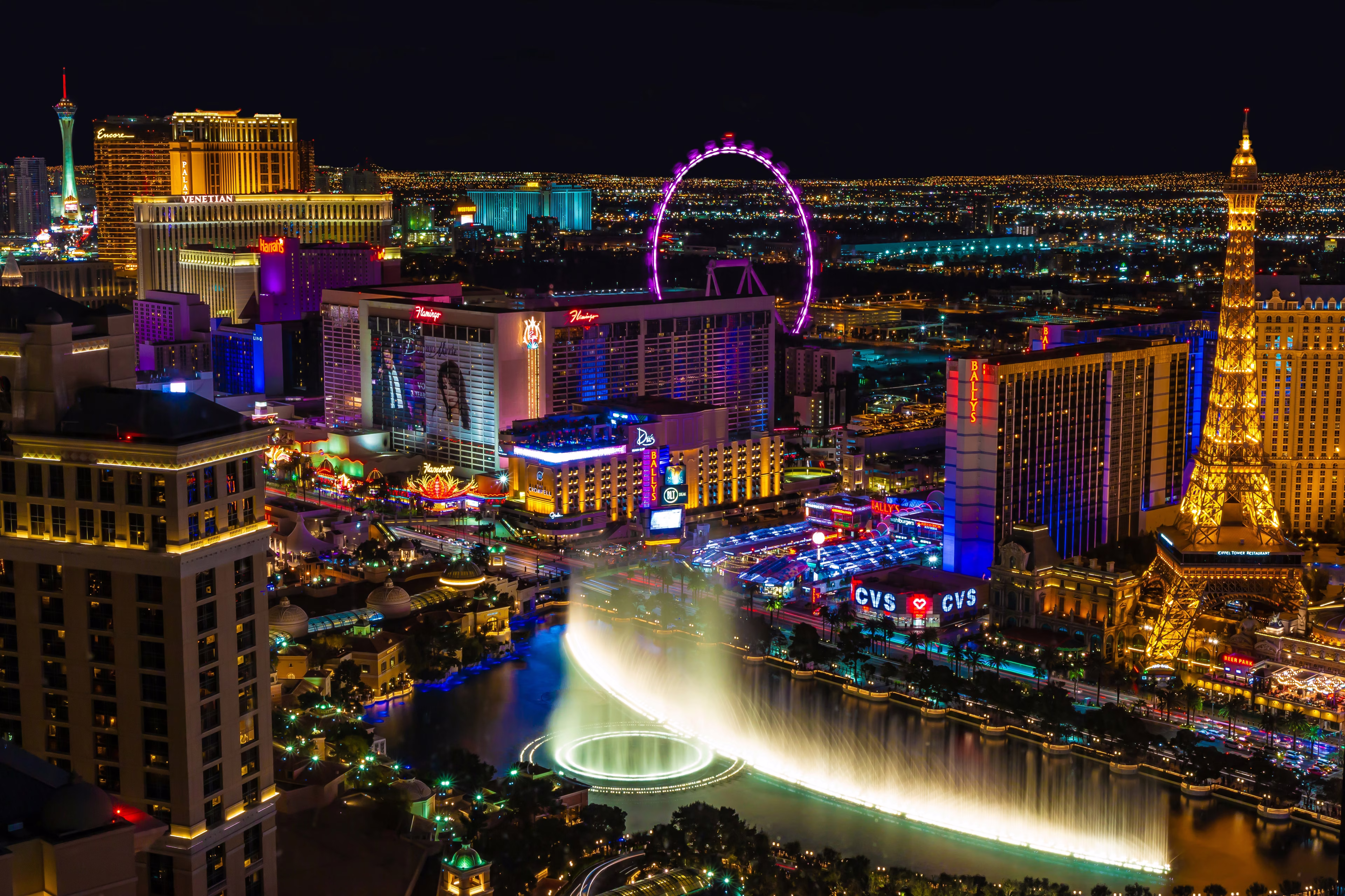 Aerial view of Las Vegas at night, featuring illuminated casinos, the Eiffel Tower replica, a Ferris wheel, and city lights.
