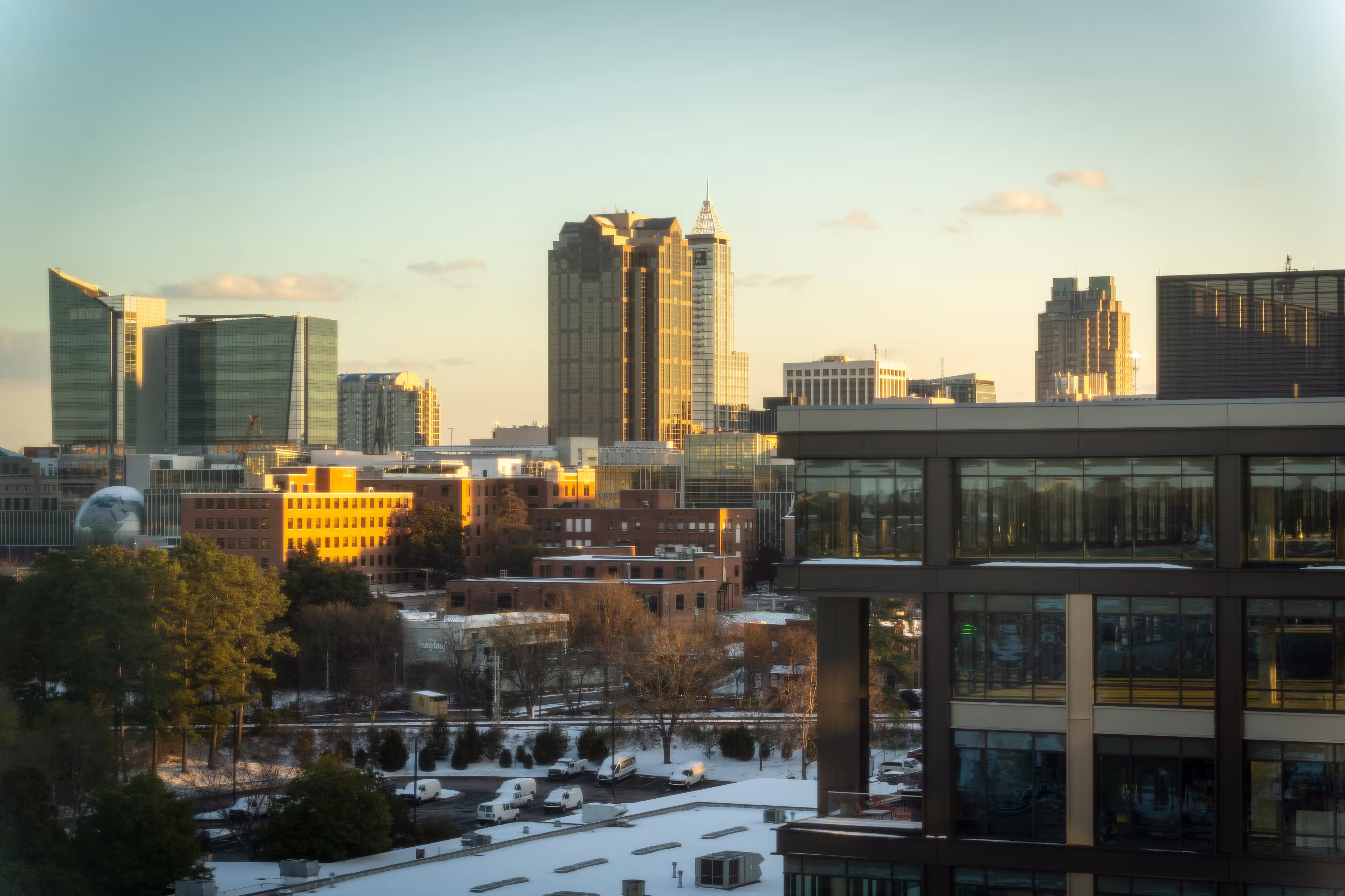 Raleigh's city skyline at sunset with modern buildings, trees, and a snow-dusted foreground.