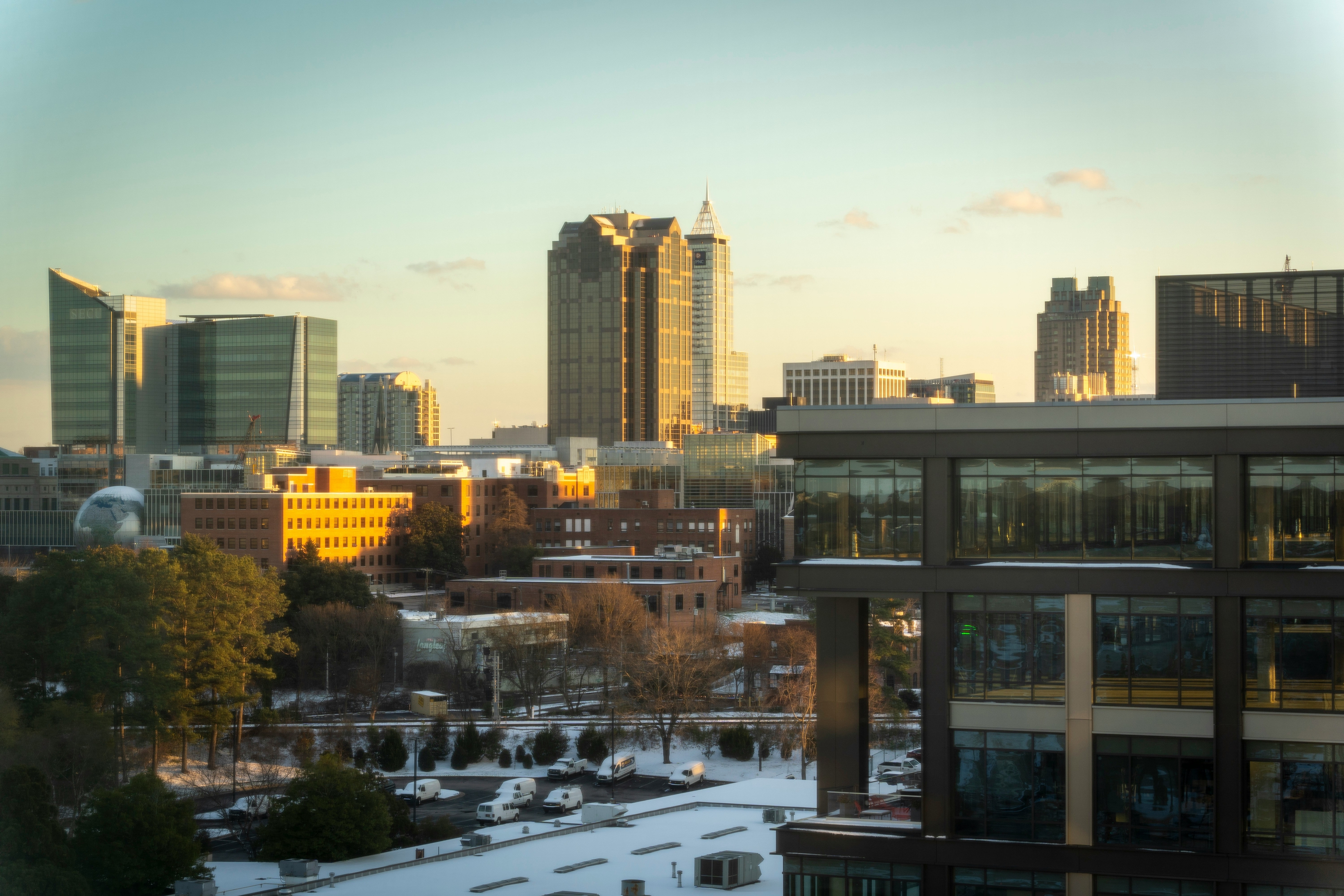 Raleigh's city skyline at sunset with modern buildings, trees, and a snow-dusted foreground.