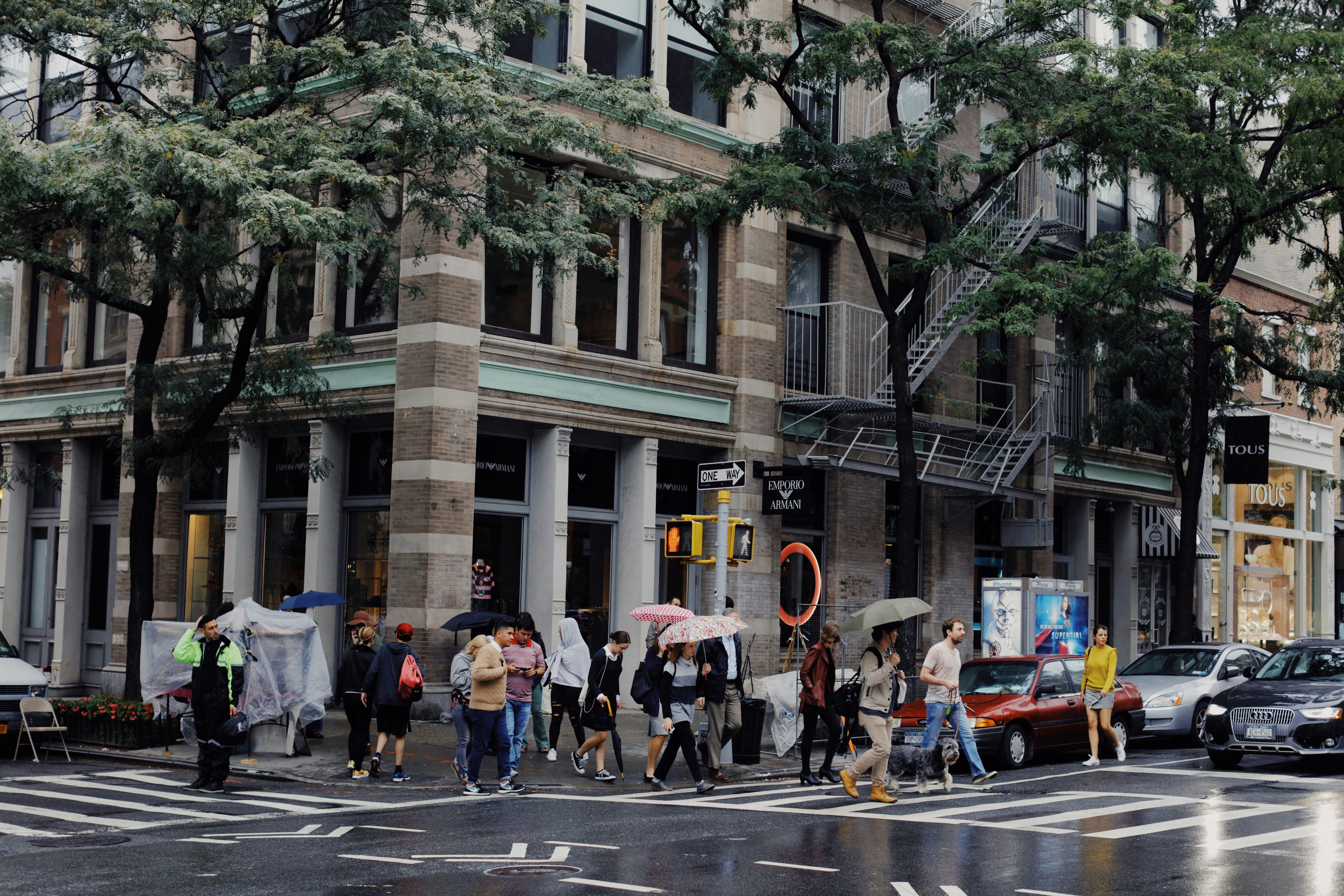 Rainy day in SoHo, New York City, with pedestrians using umbrellas while crossing in front of boutique storefronts and classic architecture.