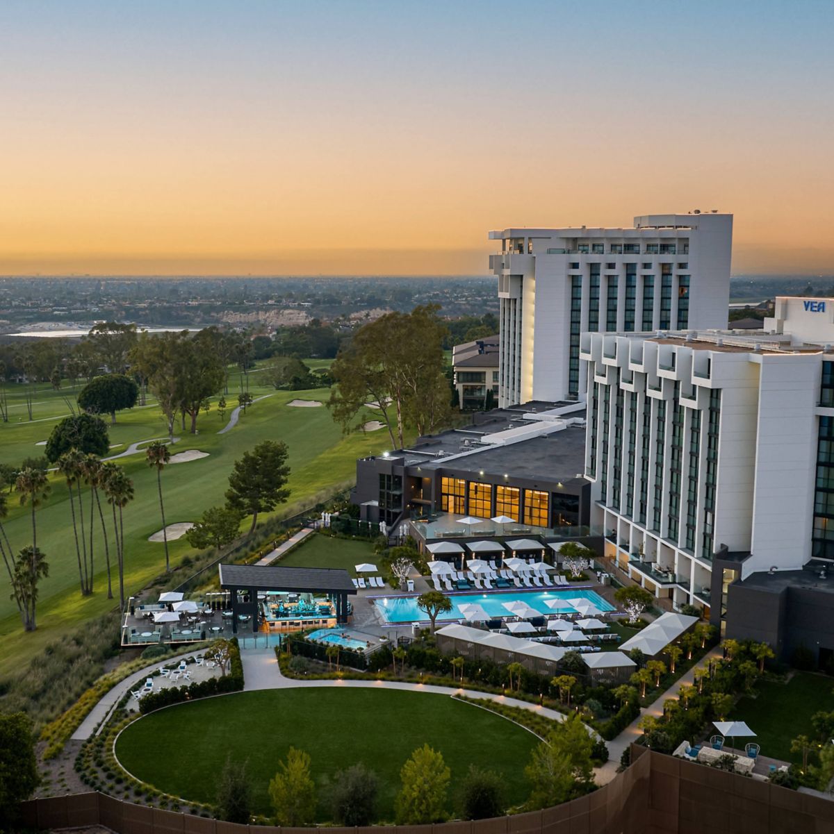 Aerial view of a luxury hotel with a pool, surrounded by greenery and a golf course, under a sunset sky.