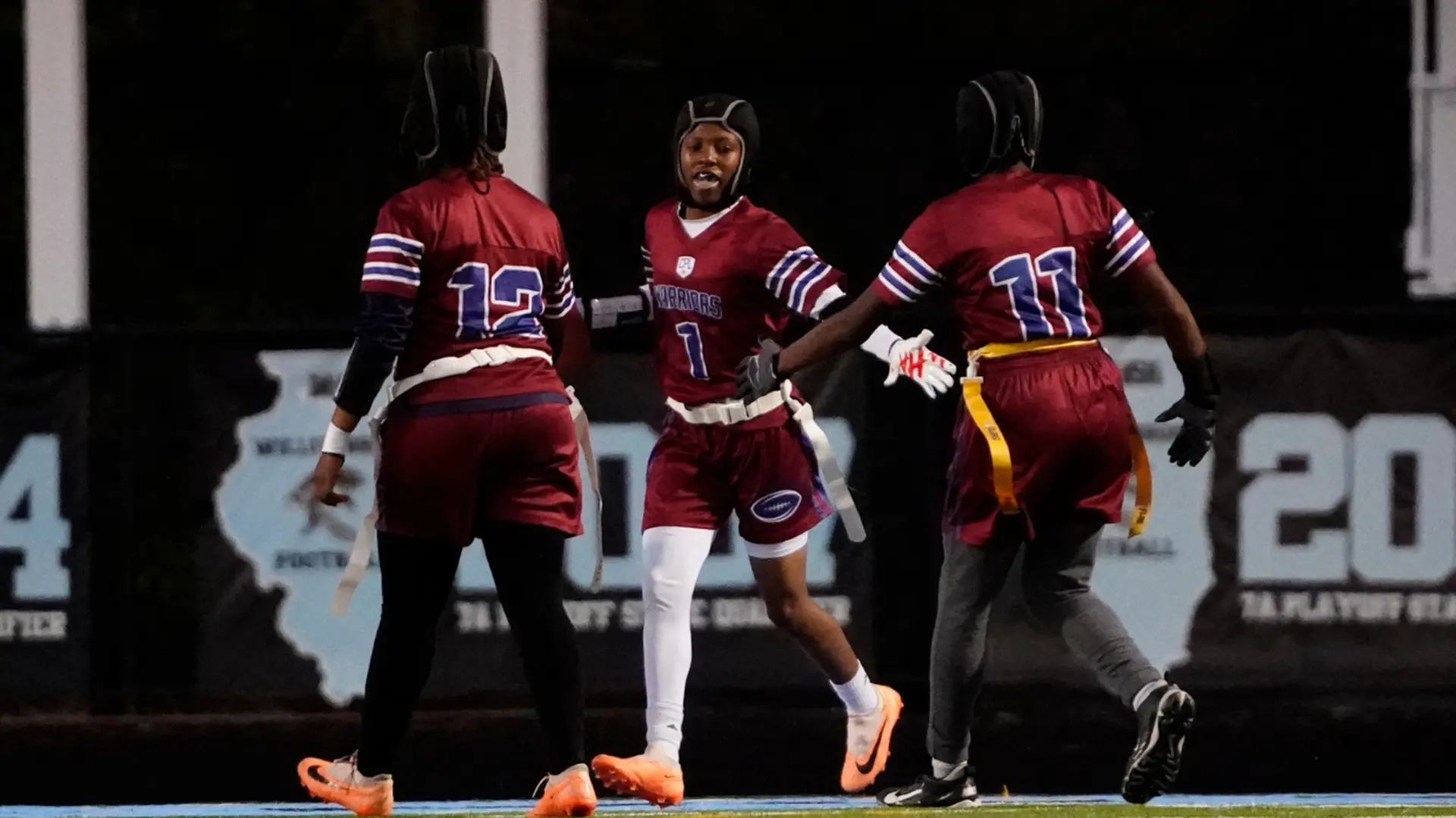 Three flag football players in maroon uniforms celebrate on a field, wearing helmets and smiling, with one player holding a football.