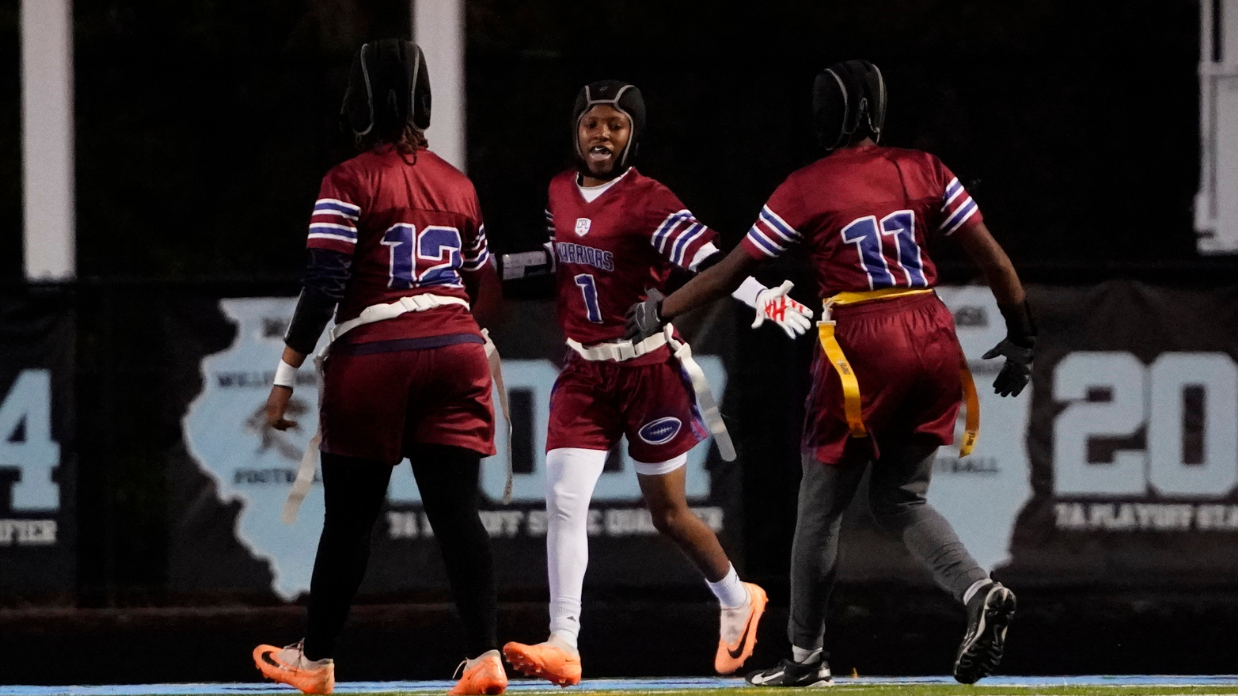Three flag football players in maroon uniforms celebrate on a field, wearing helmets and smiling, with one player holding a football.