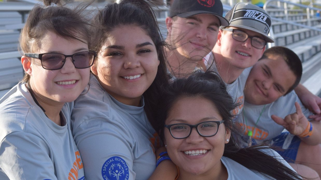 A group of smiling young adults wearing matching gray shirts, sitting closely together on bleachers in an outdoor setting.