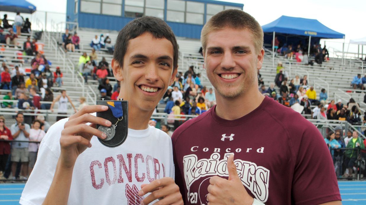 Two young men smiling, one holding a medal. Both wear "Concord" shirts at a sports event with a crowd in the background.