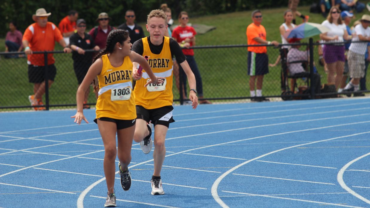 Two athletes in yellow "Murray" jerseys pass a baton during a relay race on a blue track, with spectators in the background.